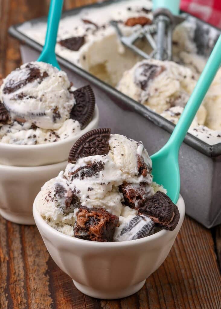 Pieces of cookies and brownies are scattered over bowls of ice cream with a green spoon.