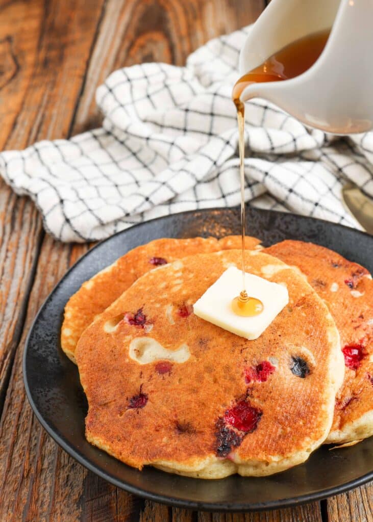 syrup pouring over cranberry filled pancakes on black plate with plaid towel