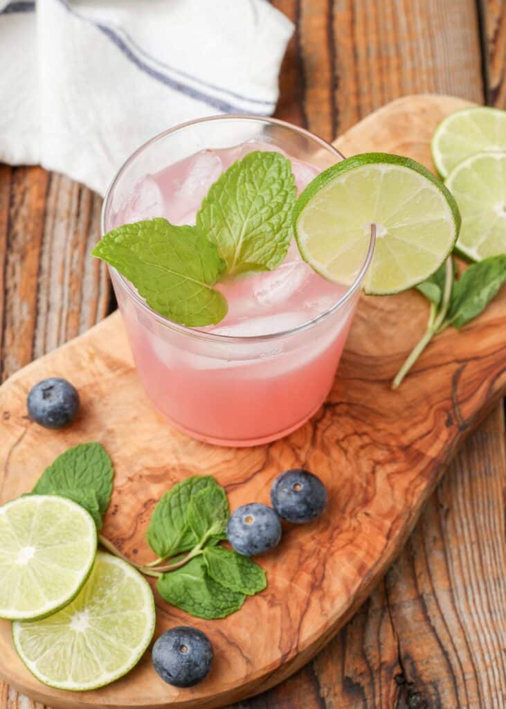 a blueberry mojito is garnished with mint leaves and a slice of lime on this cutting board, surrounded by blueberries, slices of lime, and mint leaves