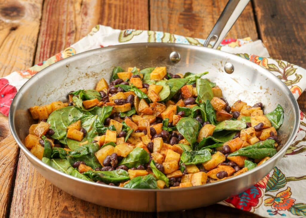 a horizontally aligned photo of a metal skillet on a wooden tabletop with a napkin in the background, the skillet contains Black Bean, Potato, Spinach Hash