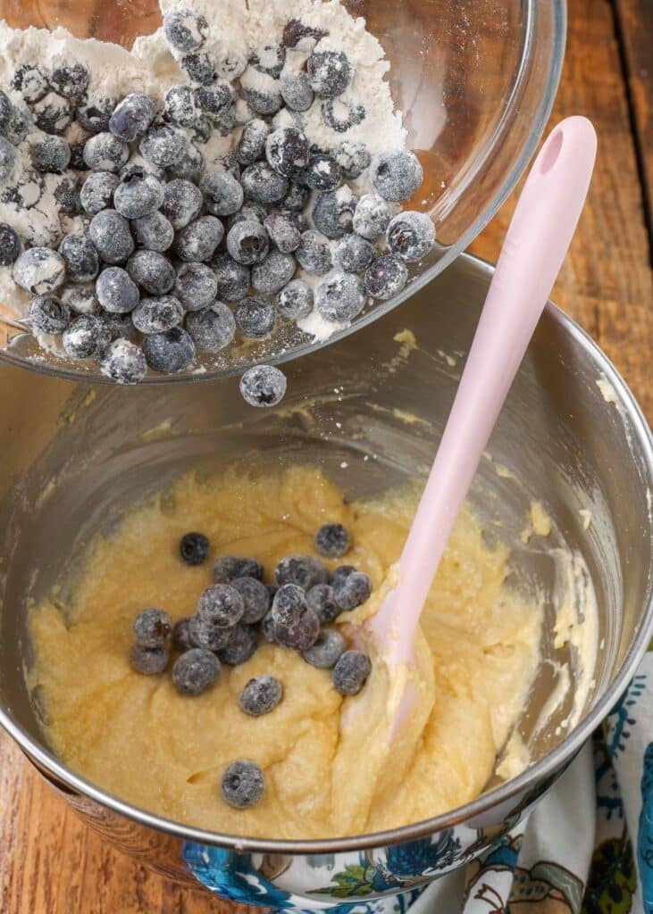 powdered blueberries being poured out of a clear glass bowl into a steel mixing bowl containing the coffee cake batter
