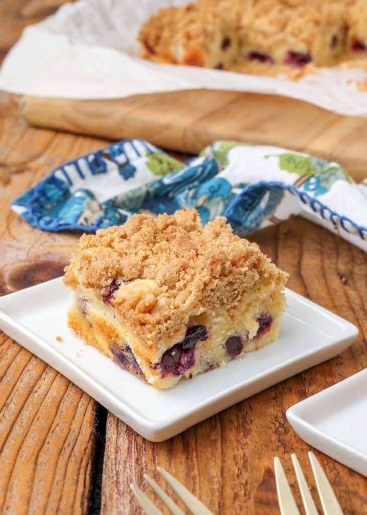 blueberry coffee cake on a white plate with a colorful blue and white towel in the background