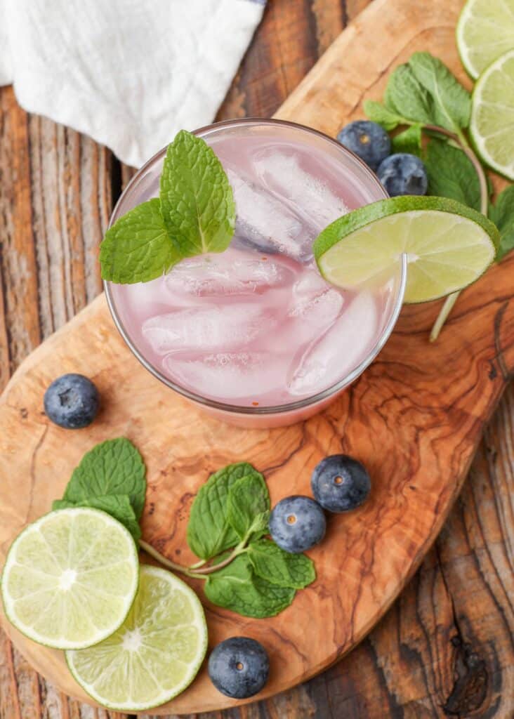 a top down shot of a blueberry mojito that has been garnished with mint leaves and a slice of lime on this cutting board, surrounded by blueberries, slices of lime, and mint leaves
