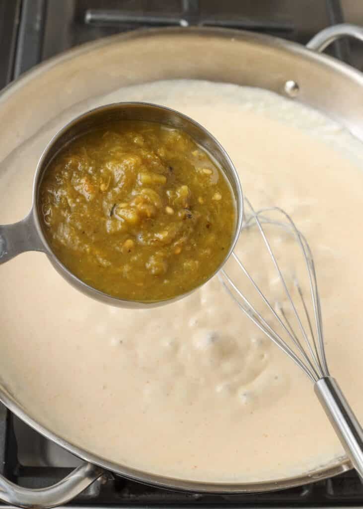 a measuring cup full of green chile is about to be added to the milk mixture in a pan with a whisk visible