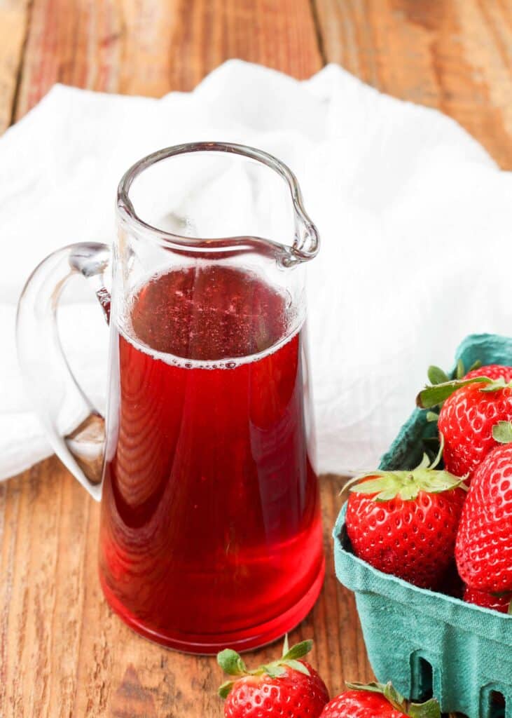 A clear pitcher containing vivid red strawberry syrup rests on a wooden tabletop with a white towel in the background