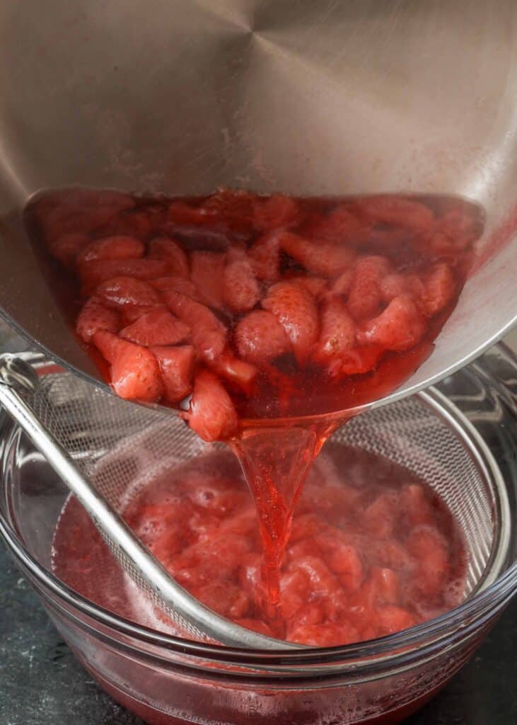 pouring cooked strawberries into the metal strainer to separate the flesh of the fruit from the juice in a clear glass bowl