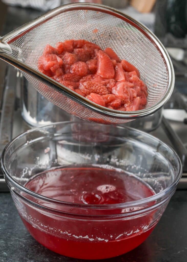 a metal strainer full of berries rests above a clear glass bowl containing the strained red juice