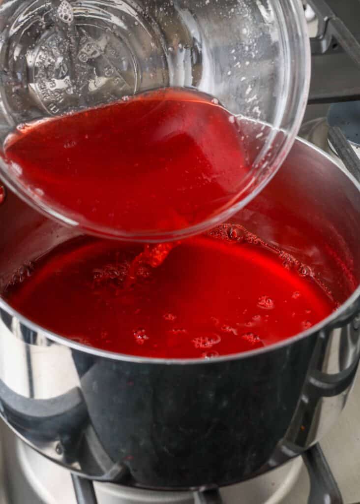 pouring the strained strawberry juice from a clear glass bowl into a metal saucepan
