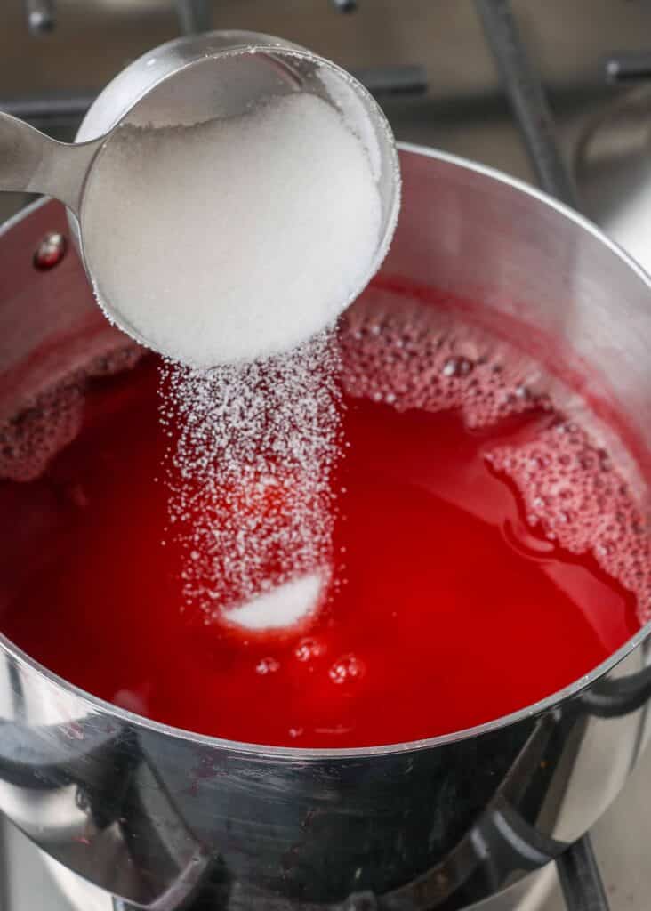 pouring sugar from a measuring cup into strawberry juice in a metal pan