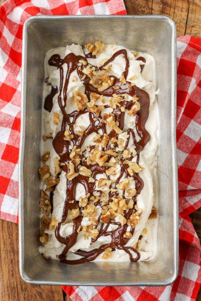 Fotografía de arriba hacia abajo de un molde para pan de metal lleno de helado de plátano rociado con chocolate y espolvoreado con nueces picadas