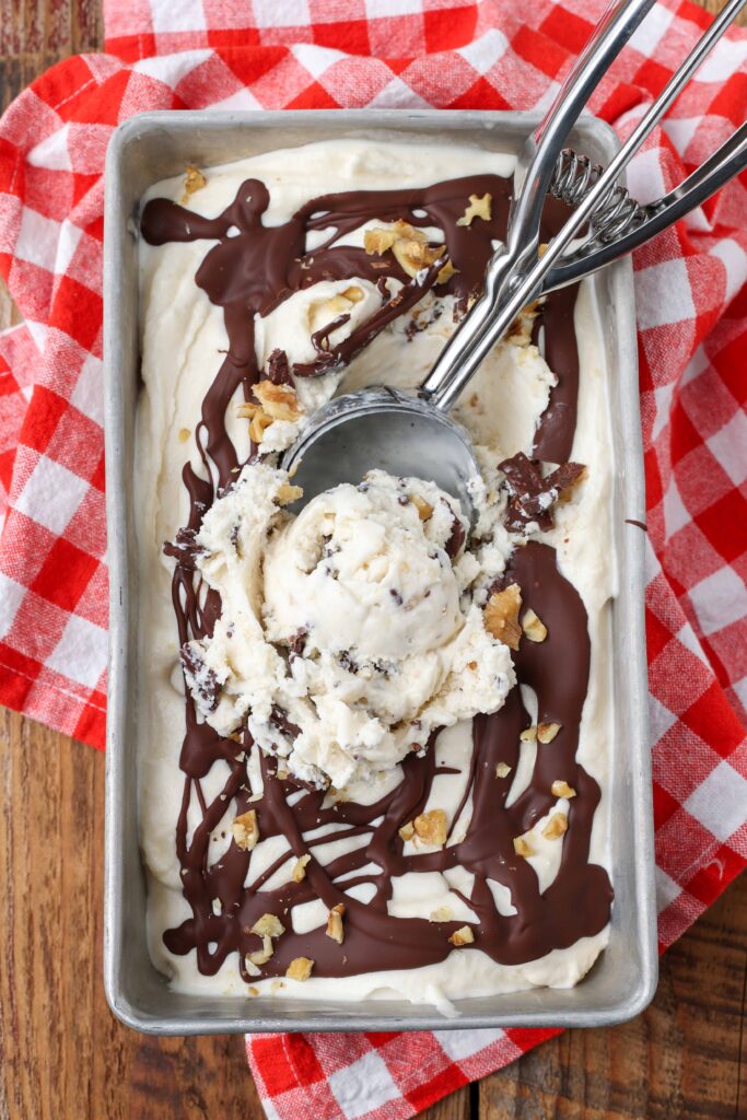 An old fashioned ice cream scoop rests in the corner of a metal pan containing banana ice cream that has been drizzled with chocolate and sprinkled with chopped walnuts