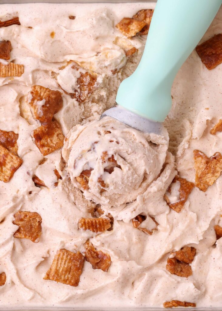 An ice cream scoop with a turquoise handle dips into a tub of cinnamon toast crunch ice cream with chunks of granola on top