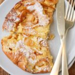 a top down photo of a white plate with part of a dutch baby with sliced apples and powdered sugar, a knife and fork resting on the plate beside the food