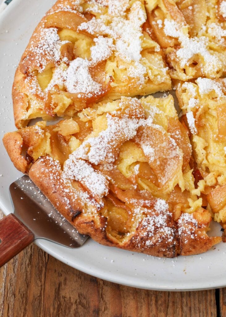 A pie server with a wooden handle lifts a wedge of german pancake off of a white plate. the german pancake has sliced apples and powdered sugar on it.