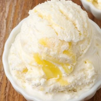 A close up shot of a scoop of vanilla ice cream swirled with lemon curd in a small white dish on a wooden table