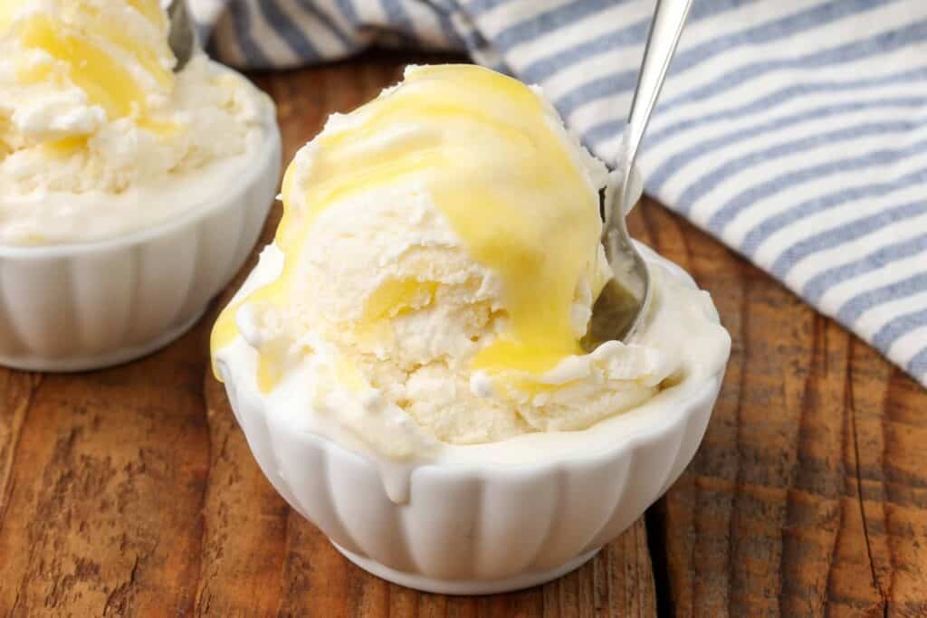 Scoops of lemon curd ice cream in a small white bowl on a wooden table against a background of blue and white striped tea towels, ice cream topped with more lemon curd
