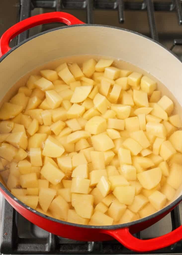 Chopped potatoes have been placed in a red handled pot on the stove to boil in water.