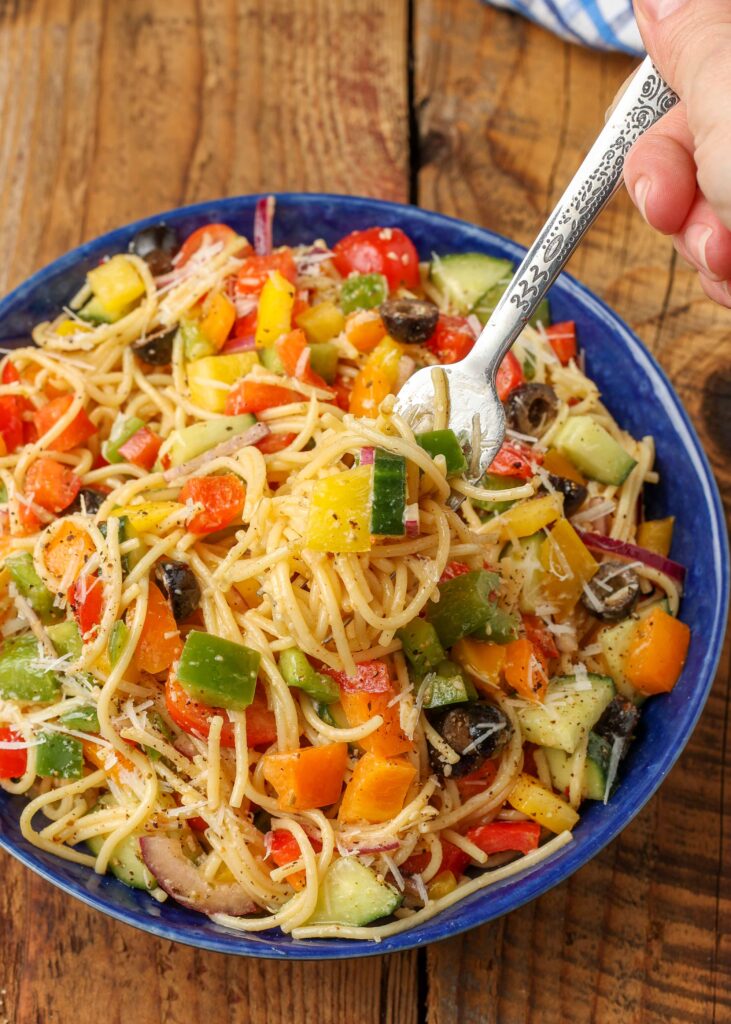 A close up of a forkful of this spaghetti salad, raised above a full blue bowl of the salad on a wooden table