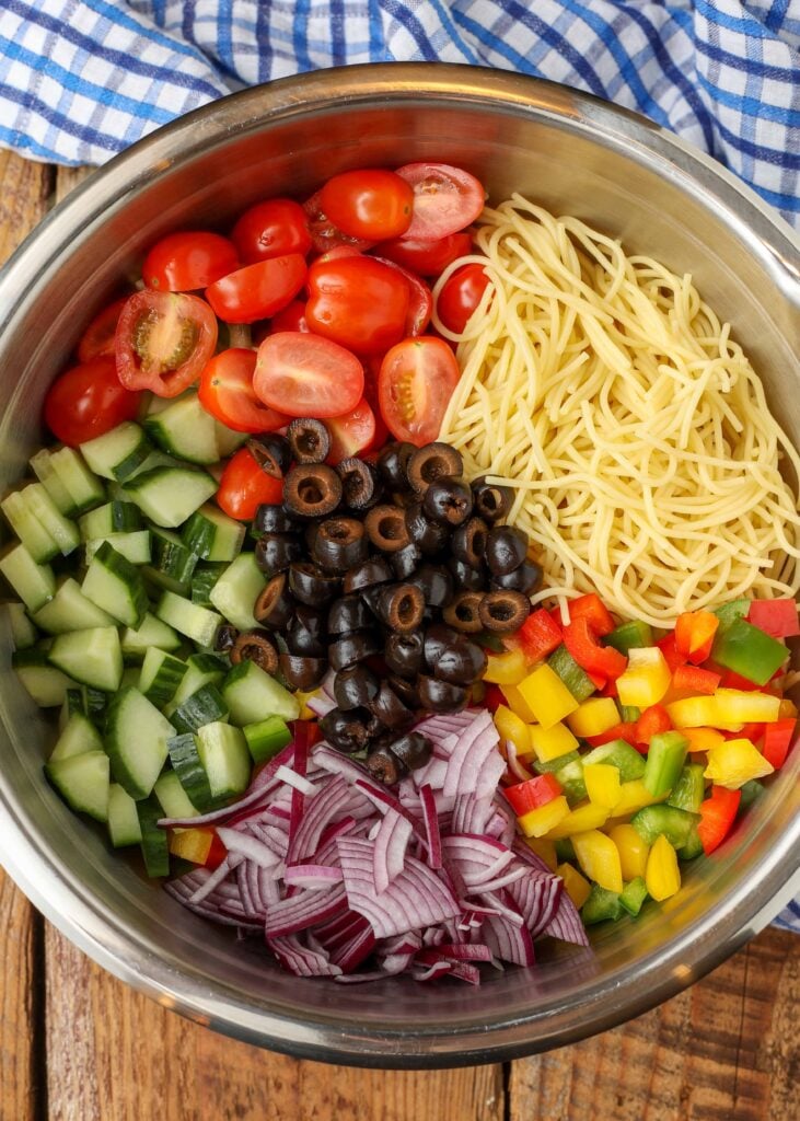 A top down shot of all the vegetable ingredients and the spaghetti noodles for this spaghetti salad in a metal mixing bowl on a wooden tabletop