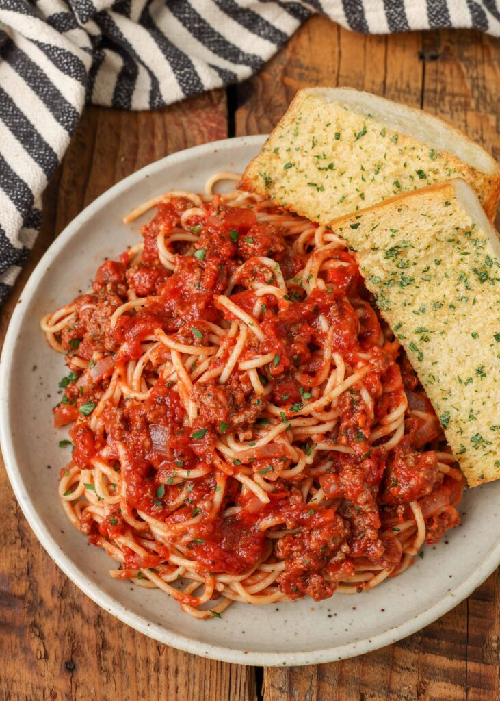 a top down photo of savory spaghetti bolognese with buttery garlic bread on a white plate on a wooden table