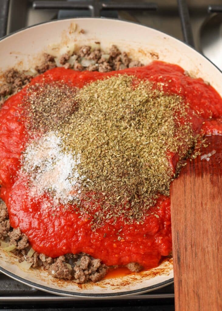 herbs and spices are piled up on top of a cherry red tomato sauce that has been poured over cooked ground beef.