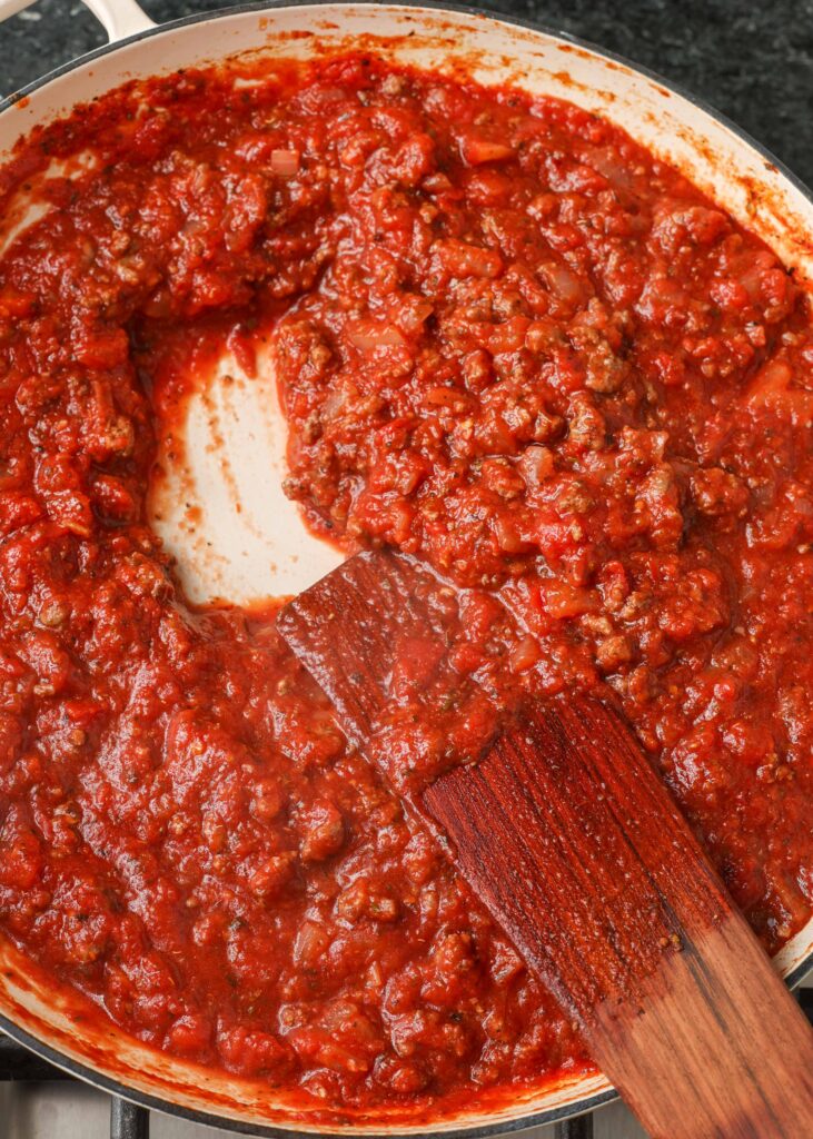 Action shot of a wooden spatula stirring spaghetti sauce in a white skillet.