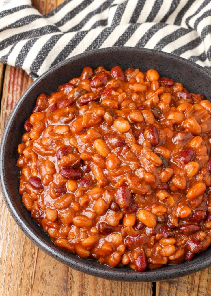 a top down image of a bowl of baked beans on a wooden tabletop with a black and white striped tea towel in the background