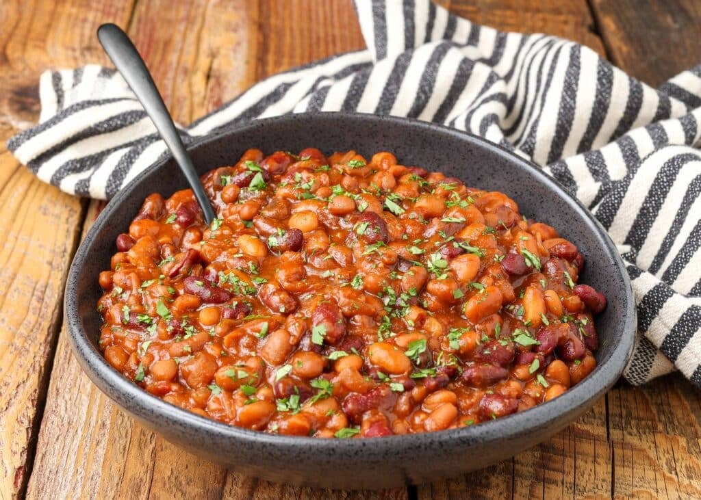 a horizontally aligned photo of a black bowl full of spicy baked beans with a black and white striped napkin in the background
