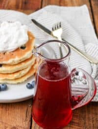 A pitcher of strawberry syrup in the forefront of this image, with a stack of blueberry pancakes in the background, topped with whipped cream