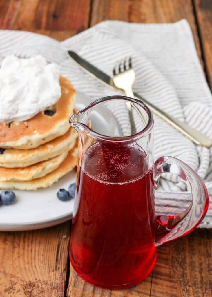 A pitcher of strawberry syrup in the forefront of this image, with a stack of blueberry pancakes in the background, topped with whipped cream