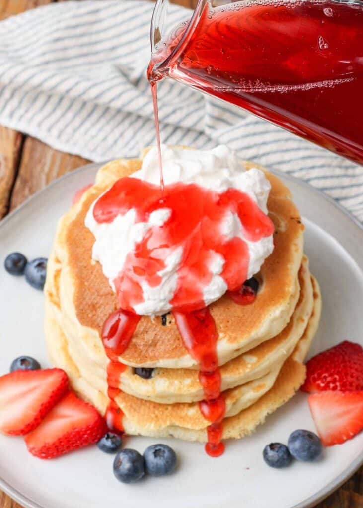 pouring strawberry syrup over a dollop of whipped cream on top of a stack of blueberry pancakes on a white plate