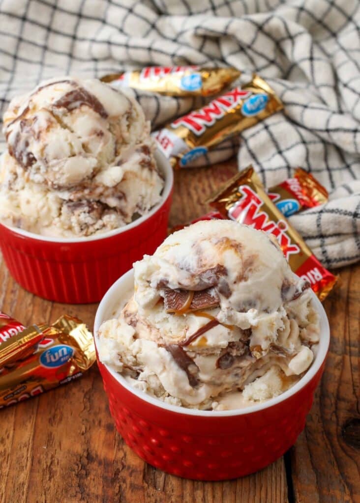 Two red ramekin with a white interior each contain a rounded scoop of ice cream with fun size twix candy bars visible in the background