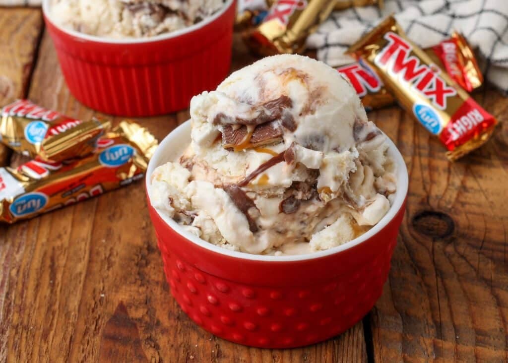 a horizontally aligned photo of a scoop of Twix Ice Cream in a red ramekin with fun size candy bars in the background
