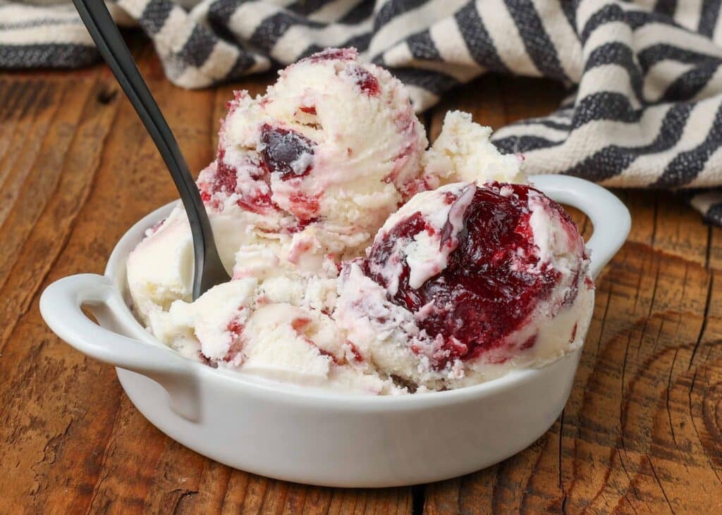 A shallow bowl filled with scoops of black cherry ice cream on a wooden tabletop with a black and white striped tea towel in the background