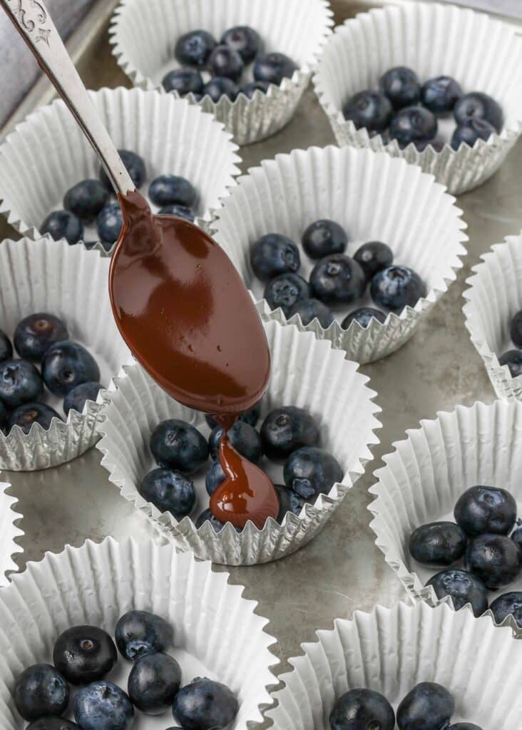 A spoonful of chocolate sauce is being poured over small clusters of blueberries organized in white cupcake wrappers