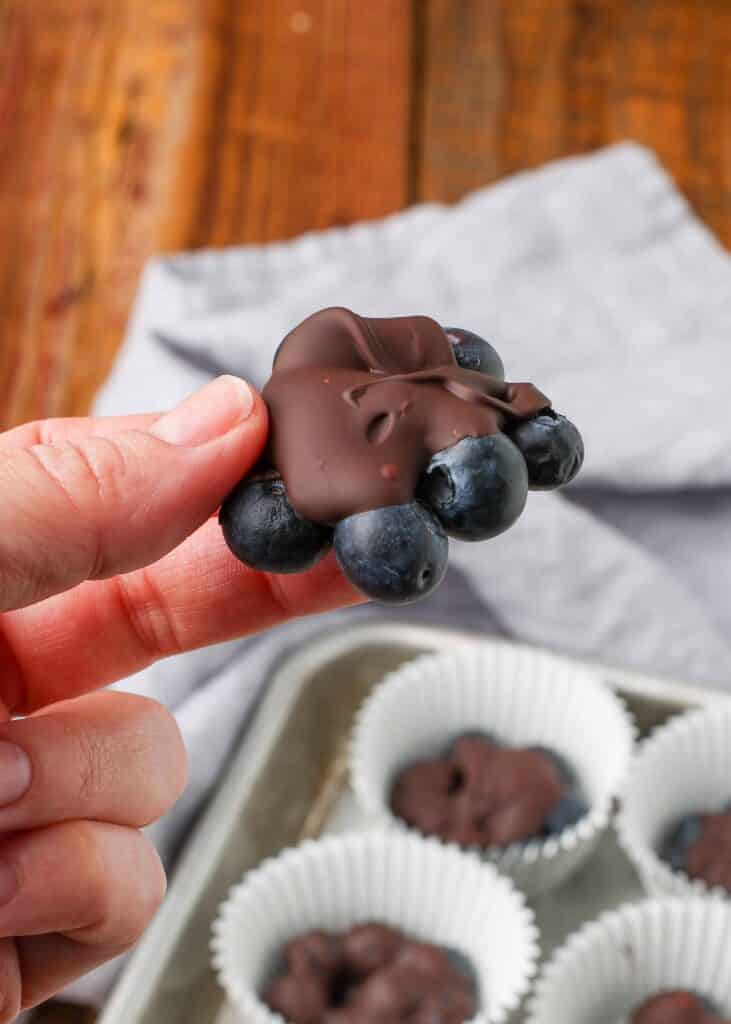 A cluster of blueberries covered in dark chocolate is being held in between a woman's thumb and forefinger