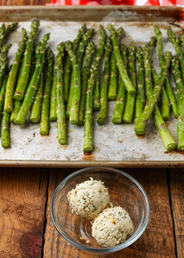 Two scoops of cowboy butter are in the trencher abreast a pan of roasted asparagus, ready to be melted.
