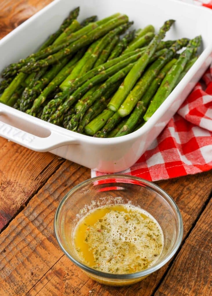 a white sultry dish is loaded with roasted asparagus spears. abreast it on a wooden tabletop there is a small glass trencher with melted garlic butter sauce.