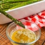 A clear glass bowl with melted compound butter in it and a spear of asparagus is being dipped in the butter.