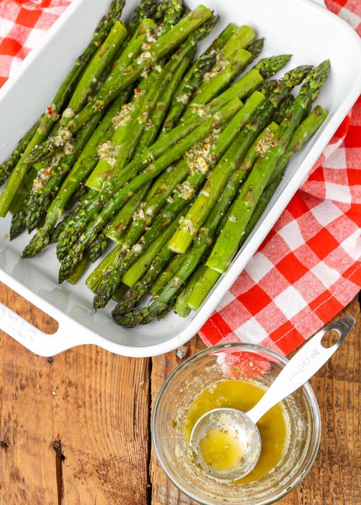 a white baking dish is loaded with roasted asparagus spears. beside it on a wooden tabletop there is a small glass bowl with melted garlic butter sauce.
