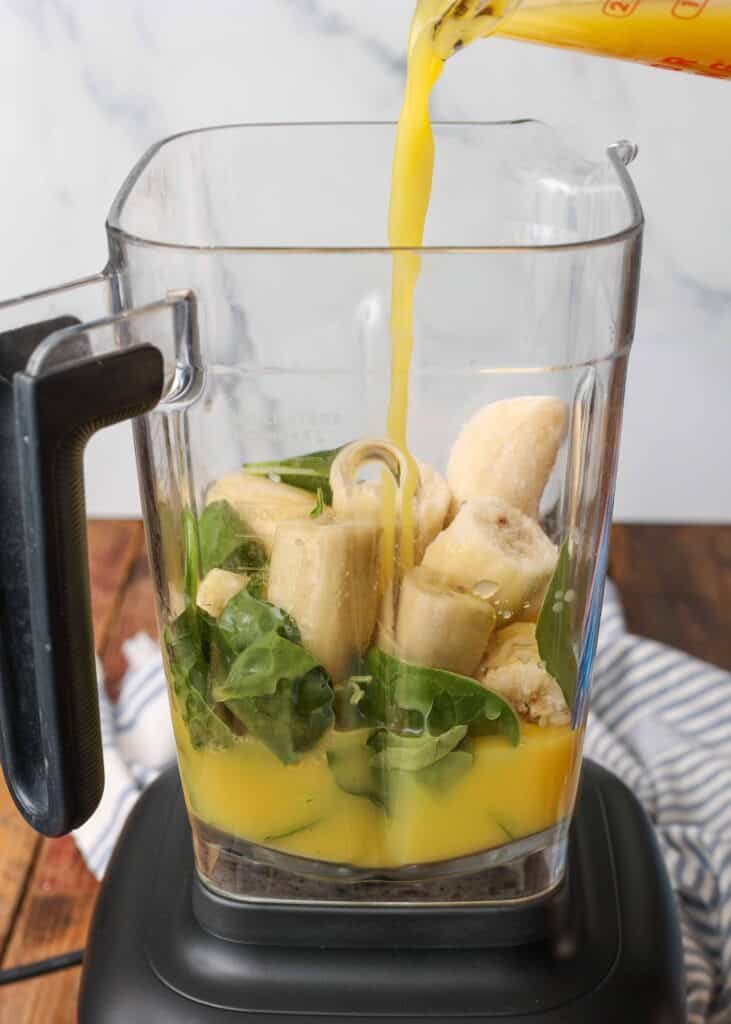 pouring orange juice over the bananas and spinach leaves in the blender
