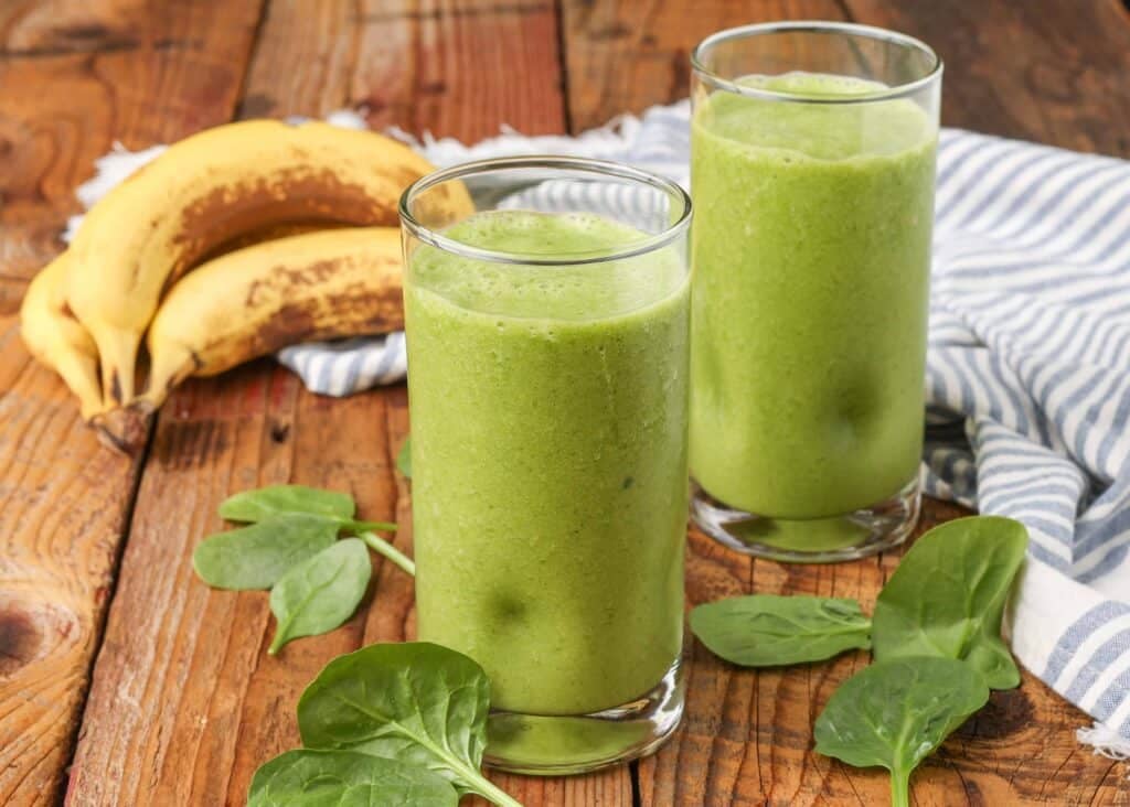 a horizontally aligned photo of spinach smoothies on a wooden tabletop with spinach and bananas scattered around them