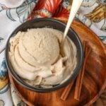 a vertically aligned image of a scoop of chai ice cream in a bowl on a wooden plate with sticks of cinnamon visible in the background