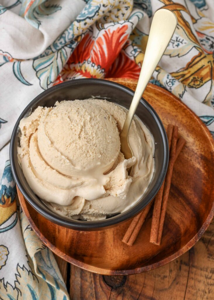 a vertically aligned image of a scoop of chai ice cream in a bowl on a wooden plate with sticks of cinnamon visible in the background