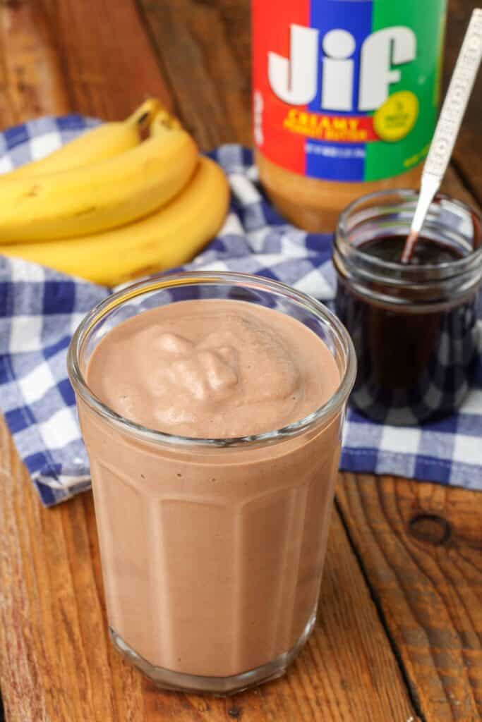 chocolate sauce, a banana, and a jar of jif peanut butter are visible in the background of this photo of a smoothie