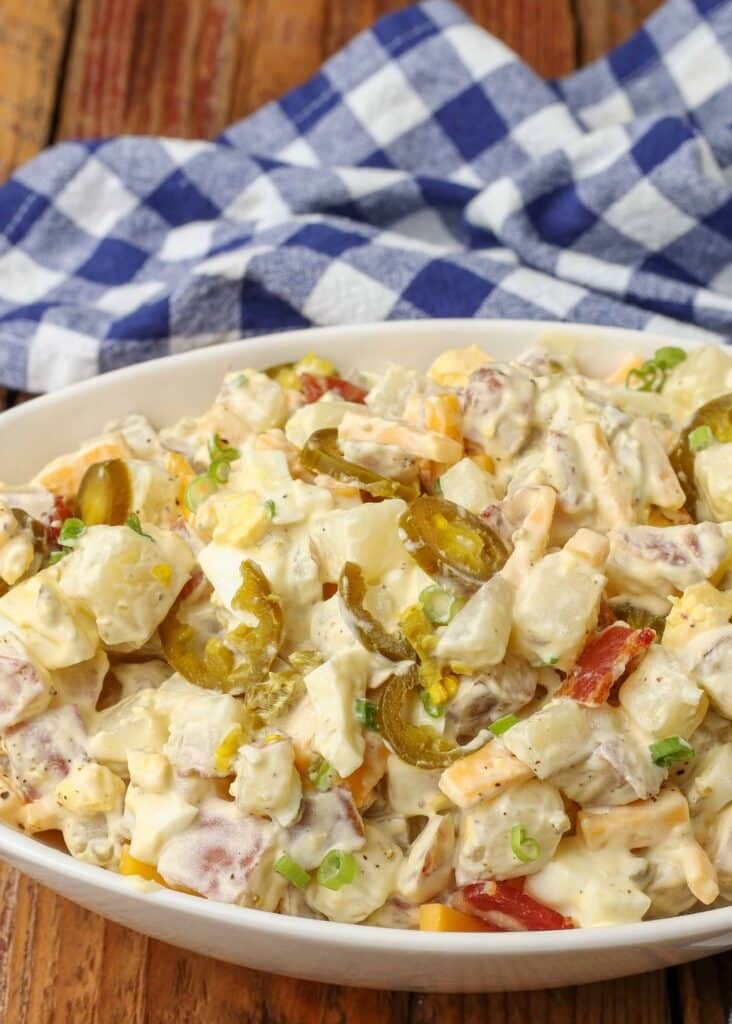 A wide shot of creamy jalapeno potato salad with bacon, cheese, green onions and jalapenos. A wide white bowl is served with a blue and white checkered towel.