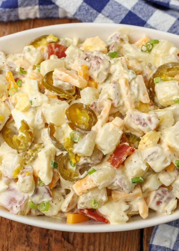 Close-up overhead shot of jalapeno creamy potato salad topped with bacon, cheese, green onions, and jalapenos, served in a wide white bowl with a blue and white checkered towel