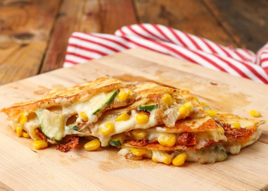 Wide shot of a stack of quartered cheesy quesadillas with zucchini and corn, served on a light brown wooden cutting board with a striped red and white towel