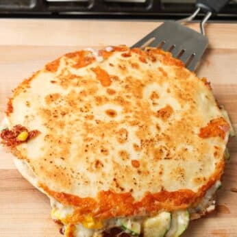 Overhead shot of double quesadilla with golden flour tortillas, green zucchini, cheese, and corn, served on a light brown wooden cutting board with a metal spatula