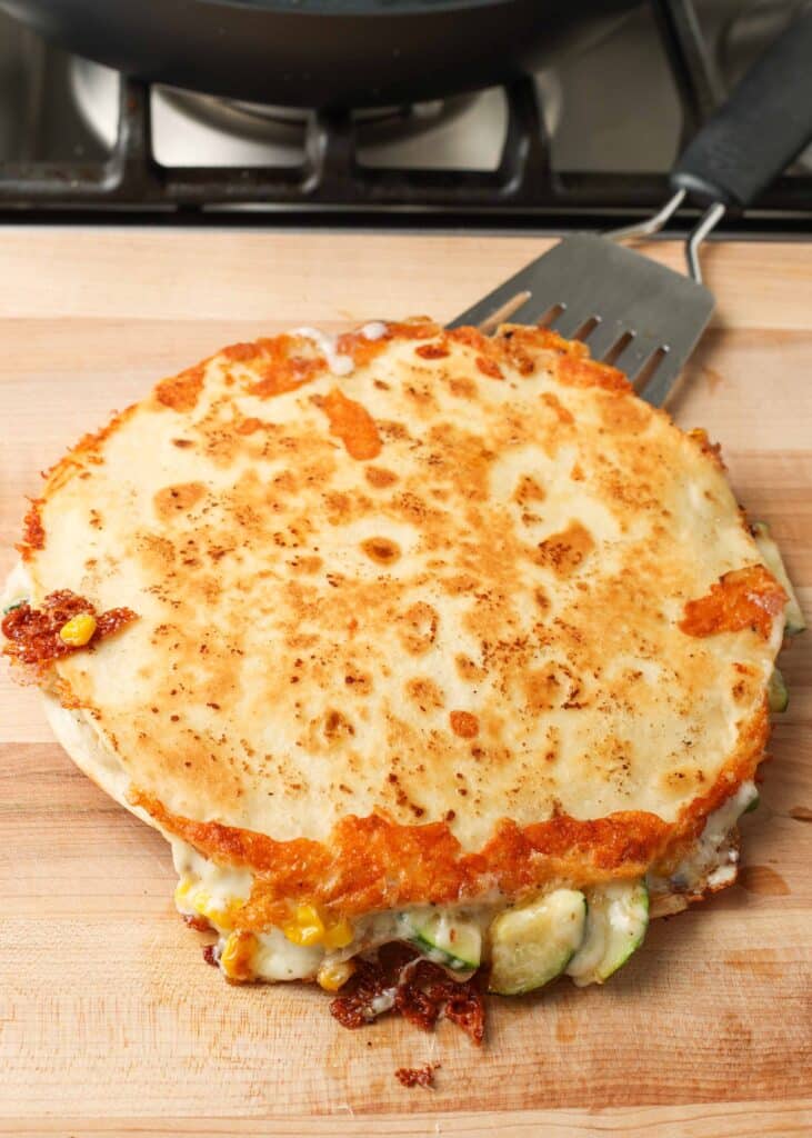 Toma superior de una quesadilla doble con tortilla de harina dorada, calabacín verde, queso y maíz, servida en una tabla de cortar de madera de color marrón claro con una espátula de metal